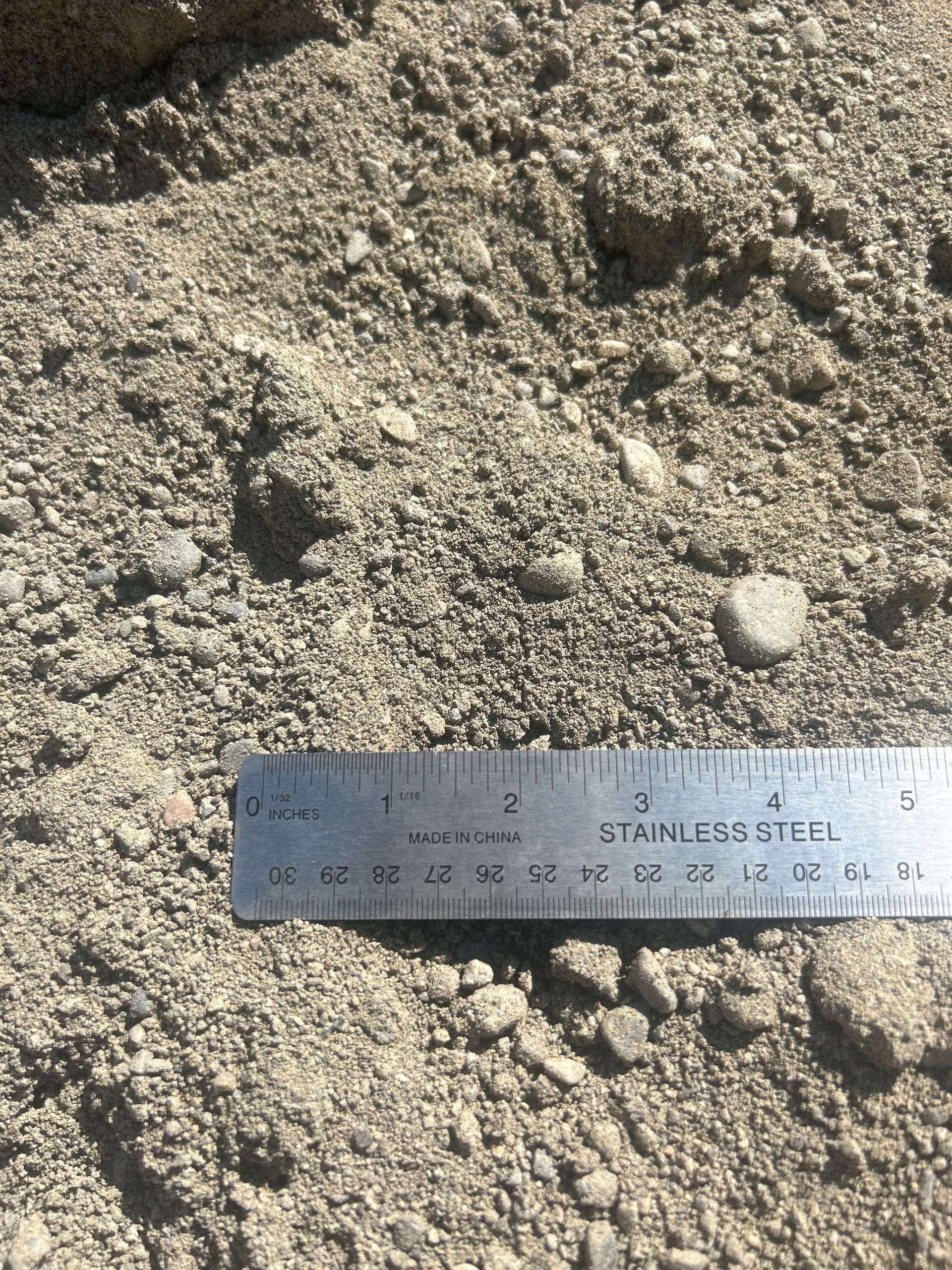 Close-up of gravel, sand, and small rocks with a stainless steel ruler for scale.