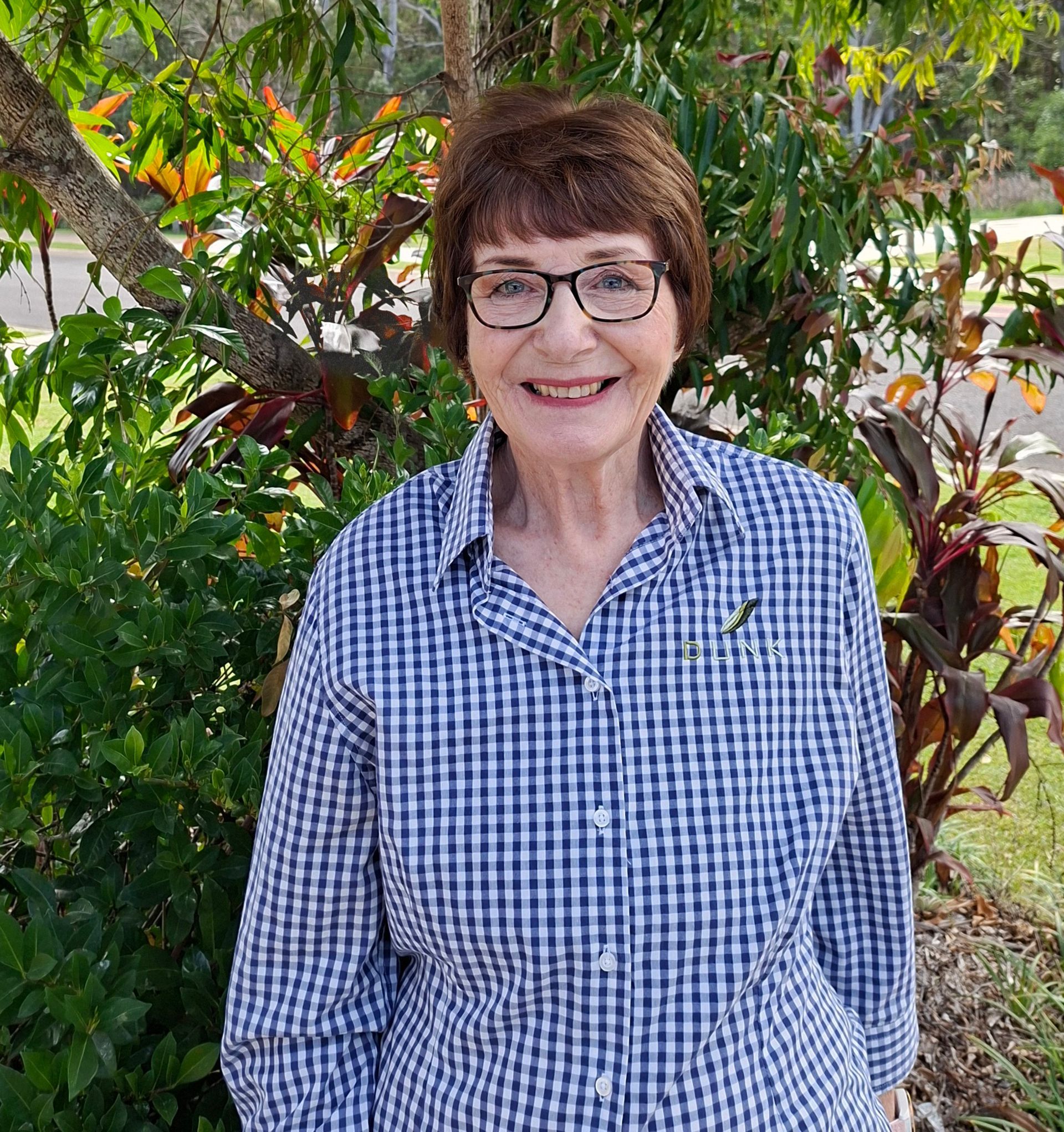 Woman in glasses and blue checkered shirt standing in a leafy garden.