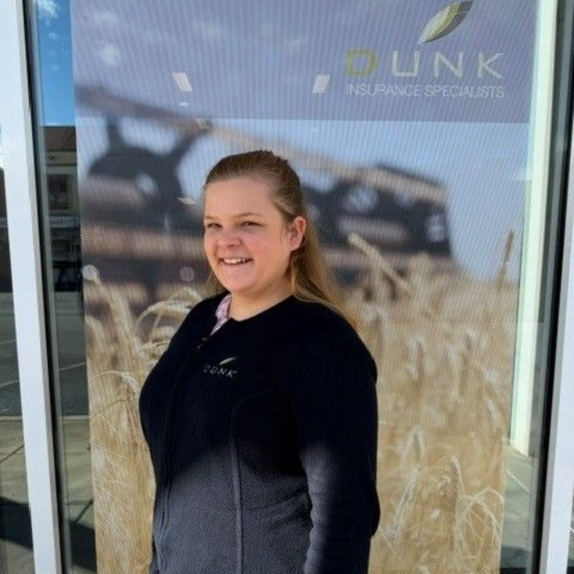 Smiling person in a black jacket standing outside a glass office window with tall grass reflection.