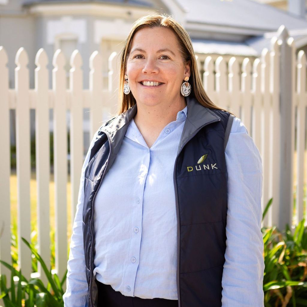 A woman wearing a blue shirt and a black vest is standing in front of a white picket fence.