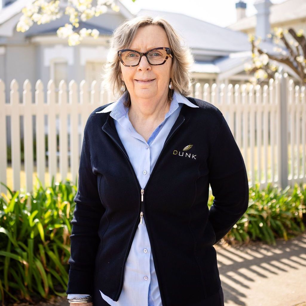 A woman wearing glasses and a black jacket is standing in front of a white picket fence.
