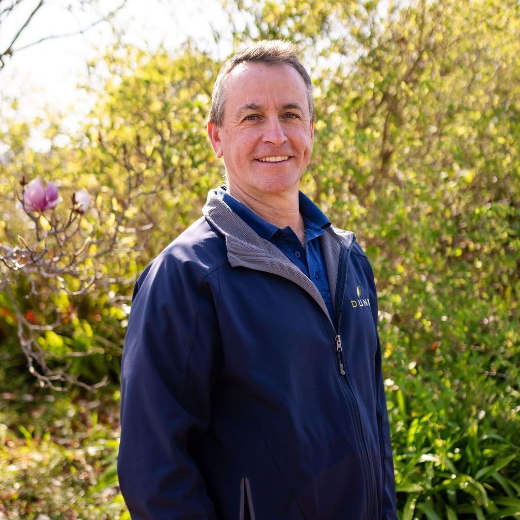 A man wearing a blue jacket and a blue shirt is standing in front of trees.