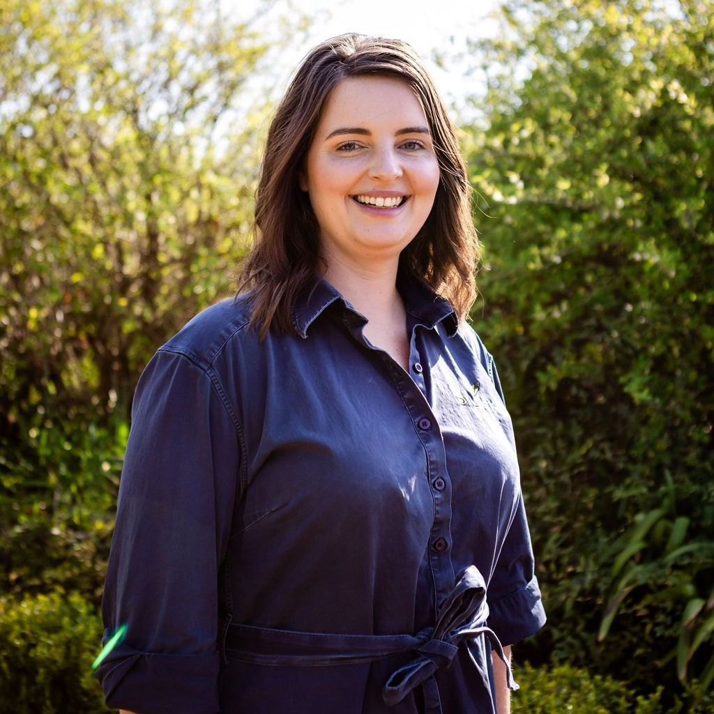 A woman in a blue shirt is smiling and standing in front of trees.