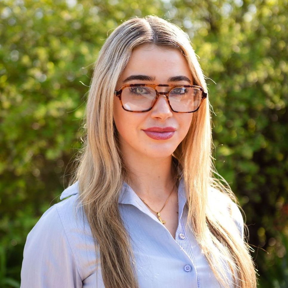 A woman wearing glasses and a blue shirt is standing in front of trees.