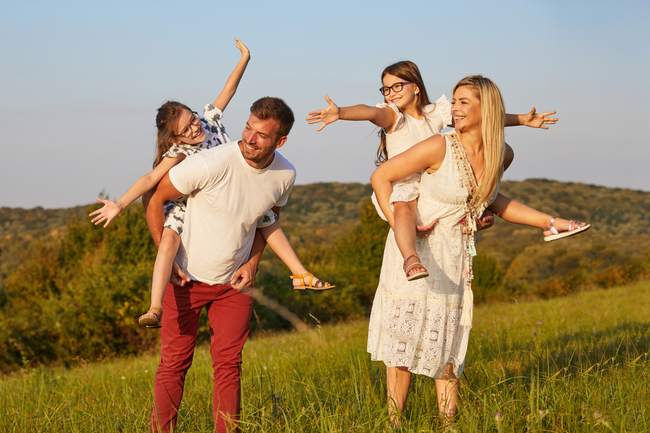 Two adults carrying two children on their backs in a sunny grassy field, smiling and playing together