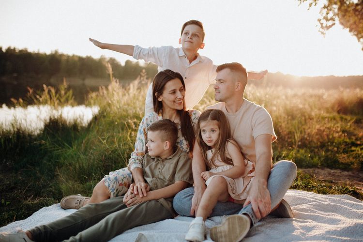 Family sitting on a blanket in a grassy field at sunset, with a man standing behind them.