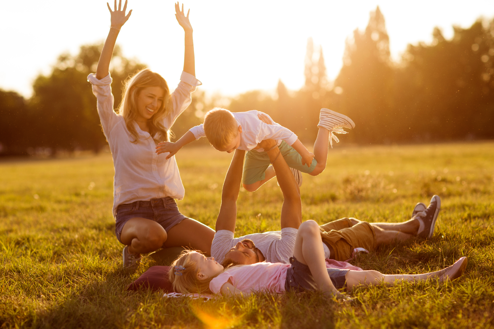 Family playing in a sunlit field, with children laughing and one being lifted into the air at sunset.