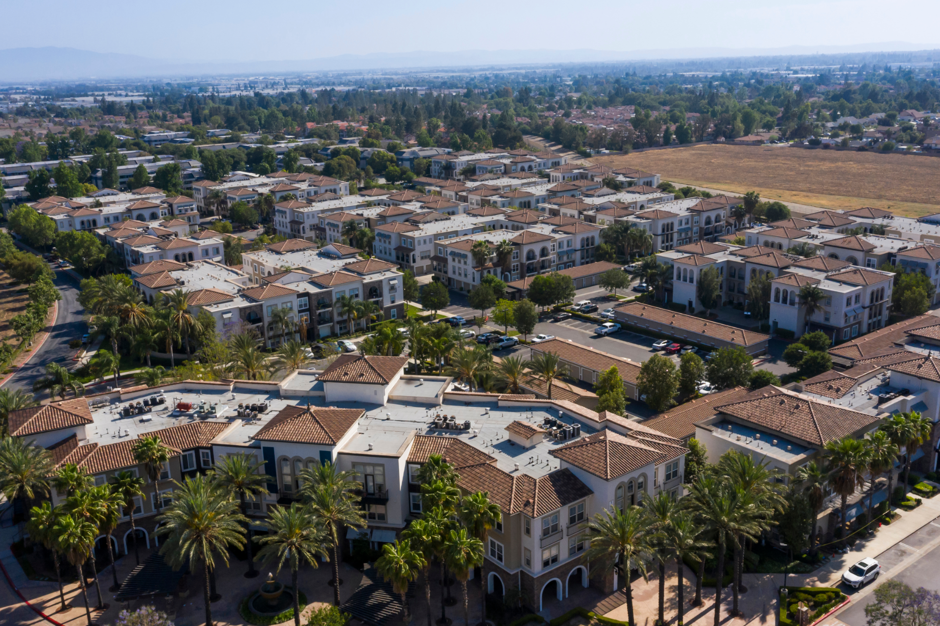 Aerial view of a suburban neighborhood with red-roofed buildings, palm trees, and open fields in the distance