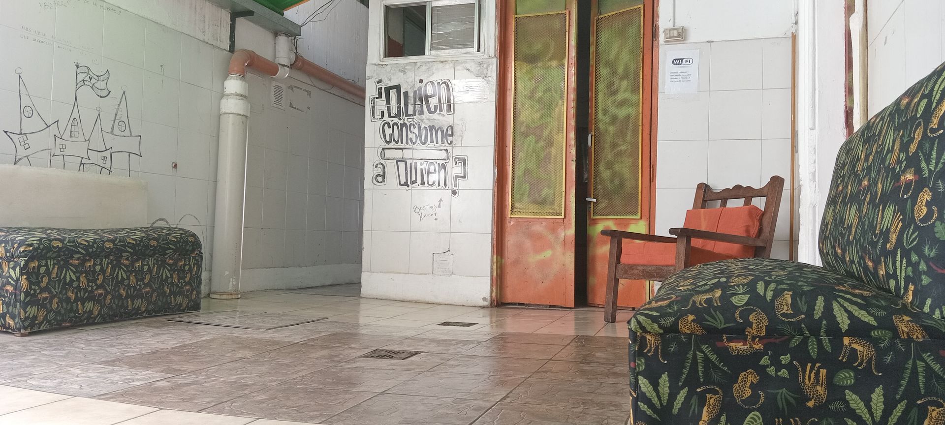 Habitación pequeña con sofás estampados, una silla de madera, paredes blancas y una puerta abierta que da a un espacio verde.
