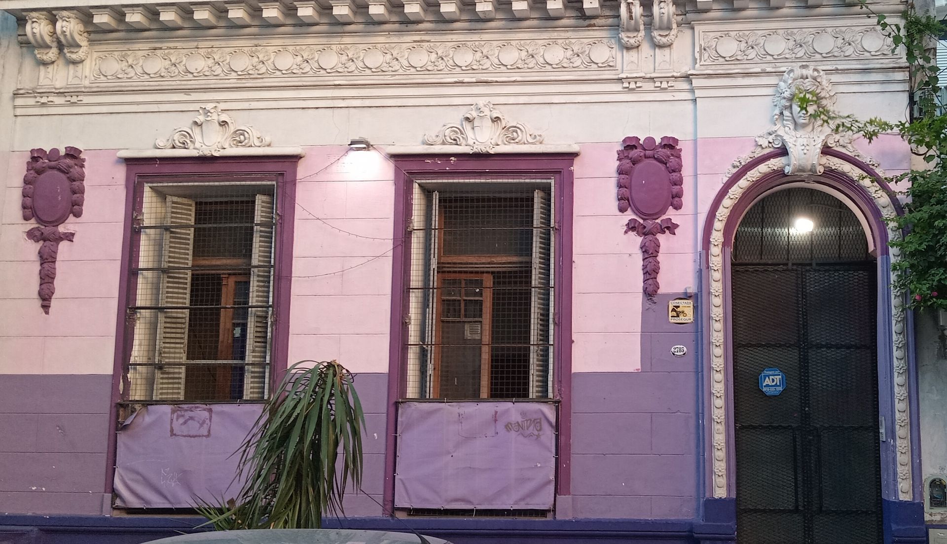 Fachada de edificio en tonos rosa pastel y lavanda con dos ventanas, una puerta y molduras decorativas blancas.