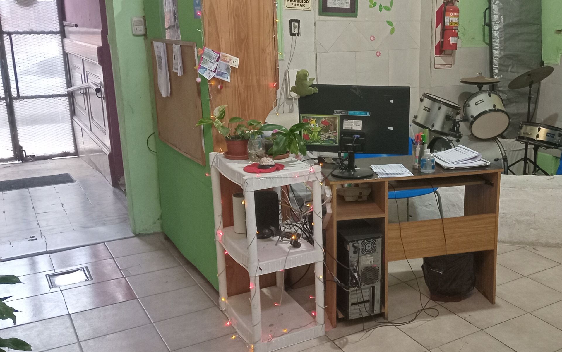 Pequeño rincón de oficina con escritorio, ordenador, estanterías y suelo de baldosas cerca de una puerta.