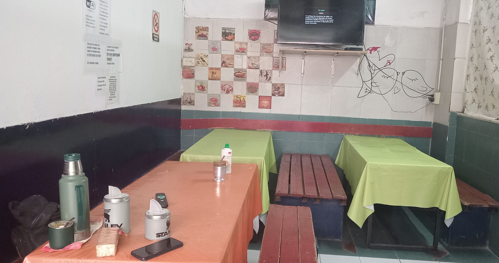 Interior de un pequeño restaurante con mesas, sillas, botellas y un televisor montado en la pared