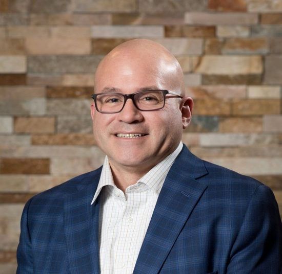 Bald man in glasses and blue plaid blazer smiles in front of a stone wall.