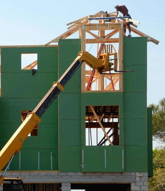 Construction worker on a roof frame, lifting wooden beams with a yellow crane, green siding.