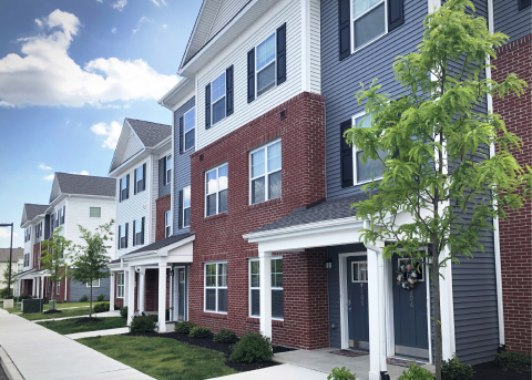 Row of multi-story townhomes with varied siding colors: white, brick, and blue. Green trees and sidewalk in front.