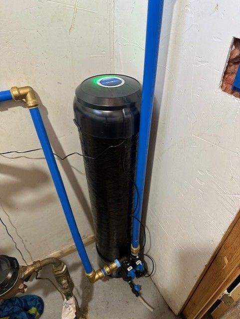 Black cylindrical water filter tank with blue pipes in a utility room corner.