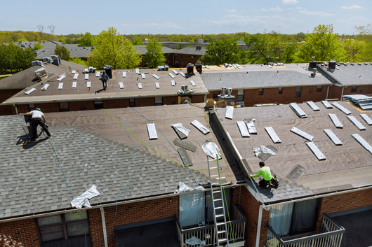 Roofers installing black shingles on a house roof, with ladders and a partly finished roof.