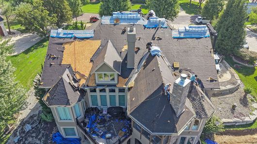 Roofers installing dark shingles on a suburban house roof with ladders and safety gear