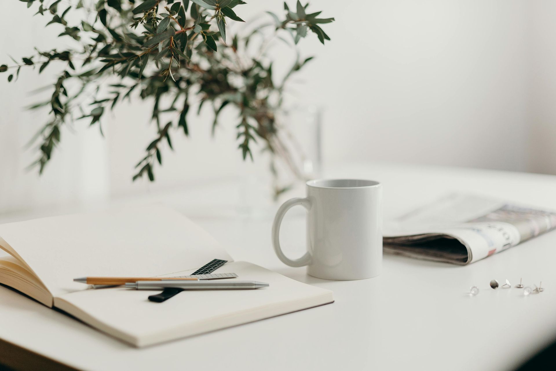 A cup of coffee , a notebook , a pen , and a newspaper are on a table.