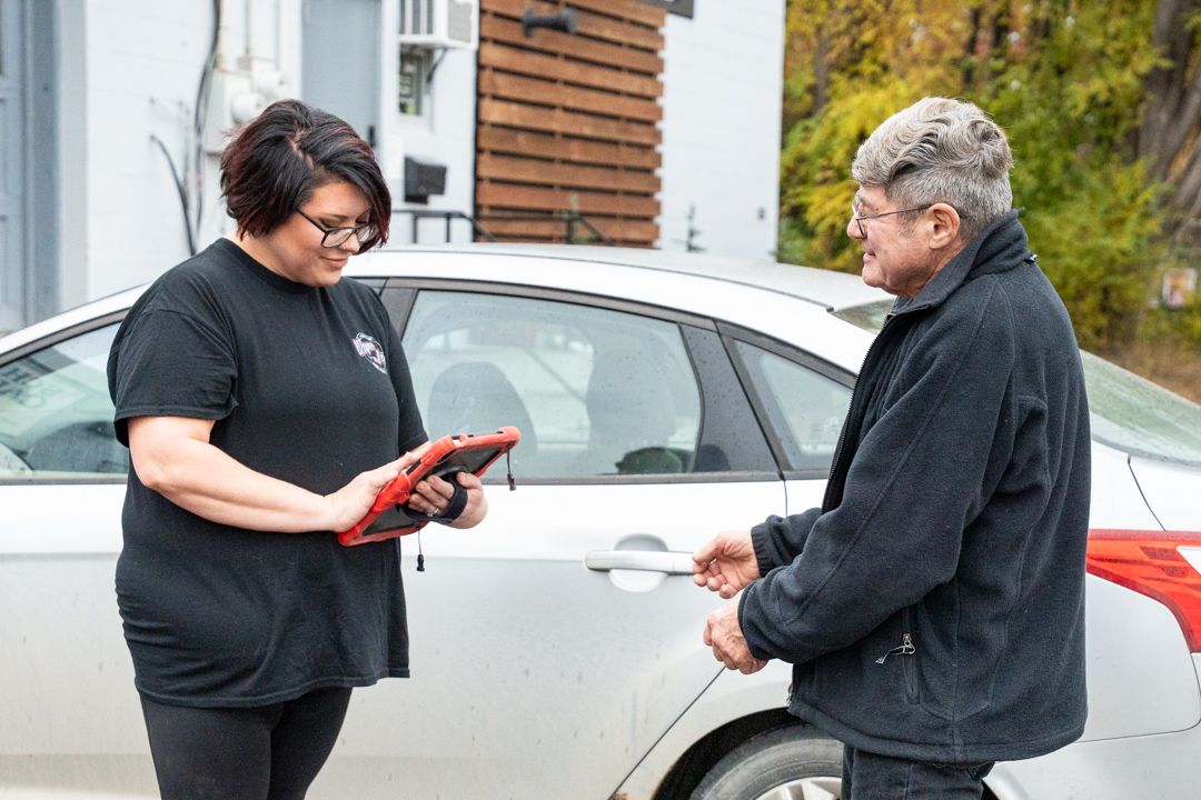 Woman in black shirt and man by silver car; woman holds tablet, man holds car door handle.
