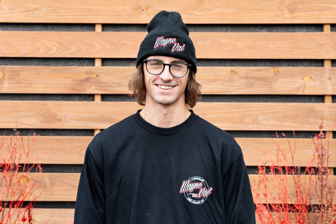 Man wearing black beanie and sweatshirt smiles in front of a wood fence.
