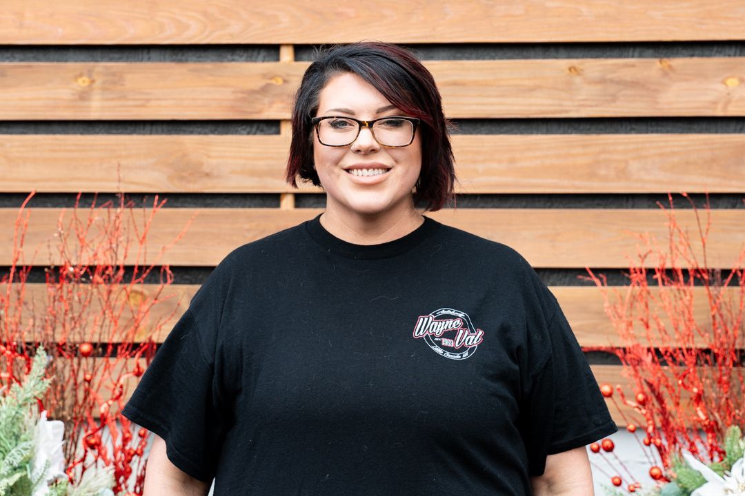Woman wearing glasses and a black t-shirt smiles in front of a wood background with red decorative branches.