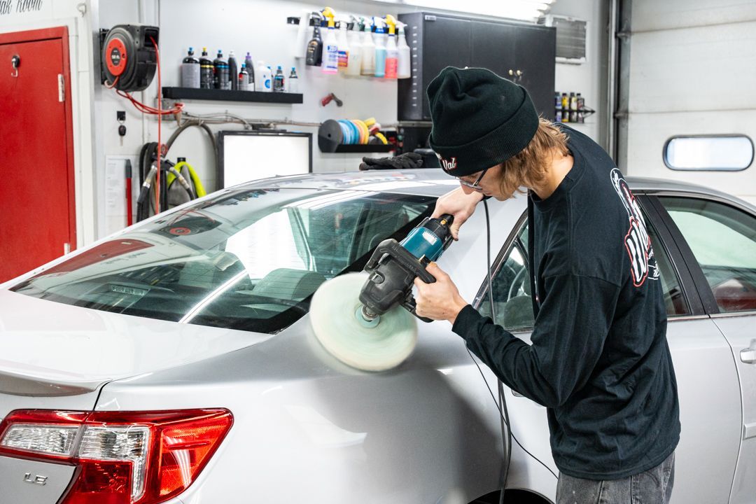 Person polishing a silver car window with a power buffer in a garage.