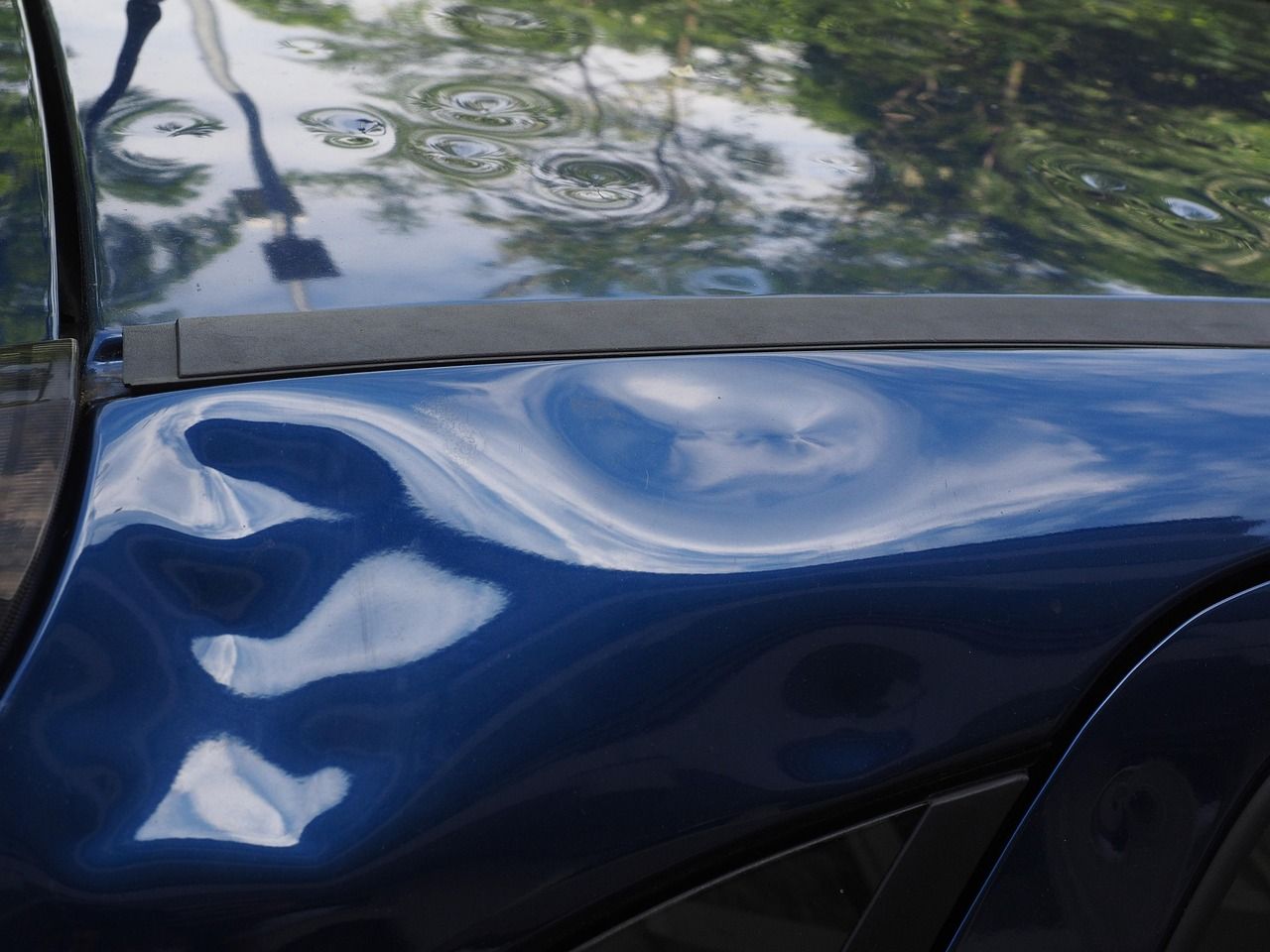 Blue car fender with several dents, reflecting sky and trees.
