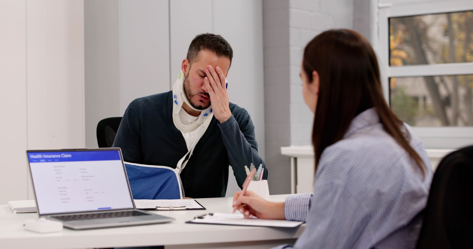 Man in a meeting rubs his face while two colleagues sit across a table with a laptop and papers.