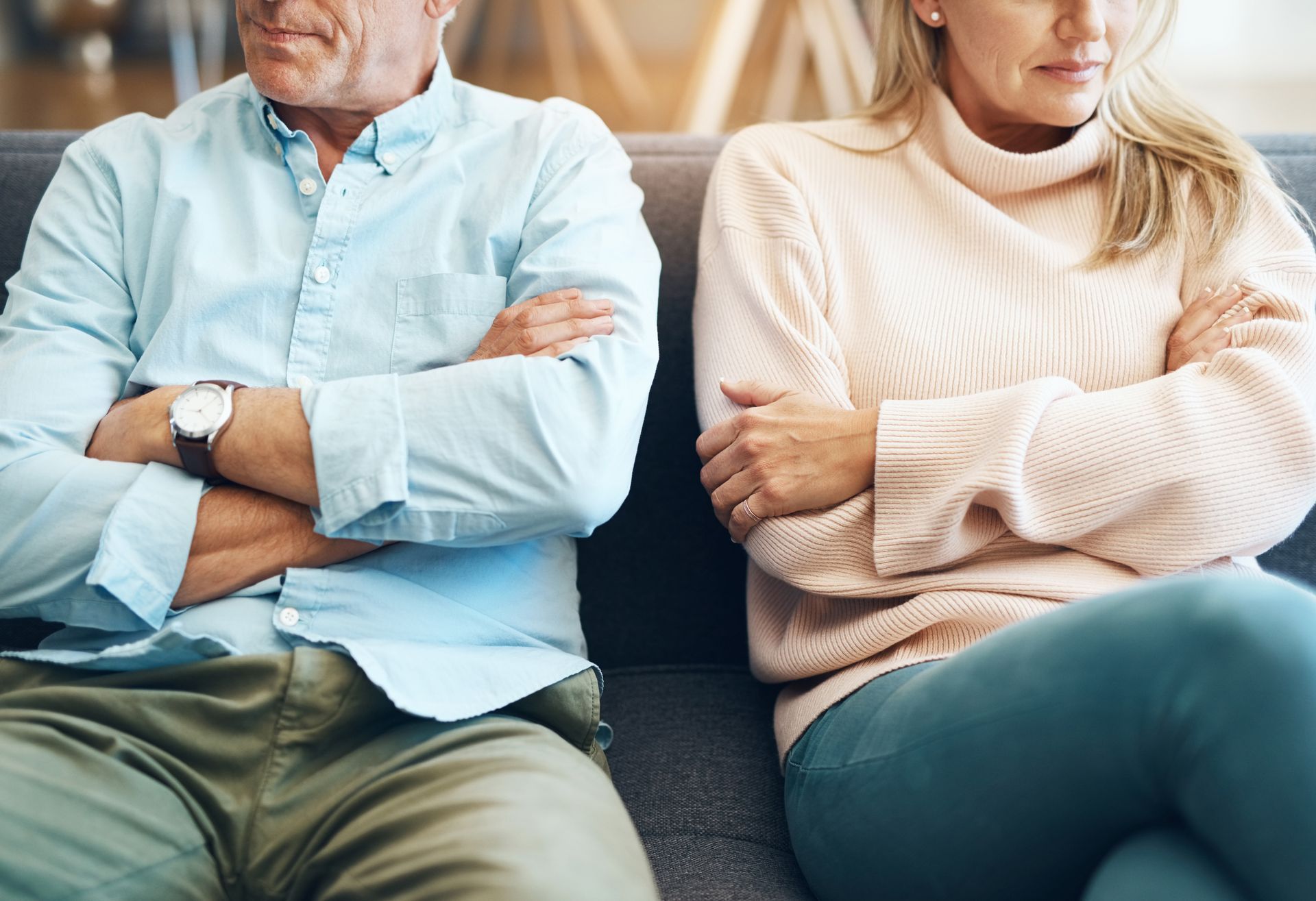 Two adults sitting on a couch with arms crossed, wearing light sweaters and shirts in a cozy indoor setting