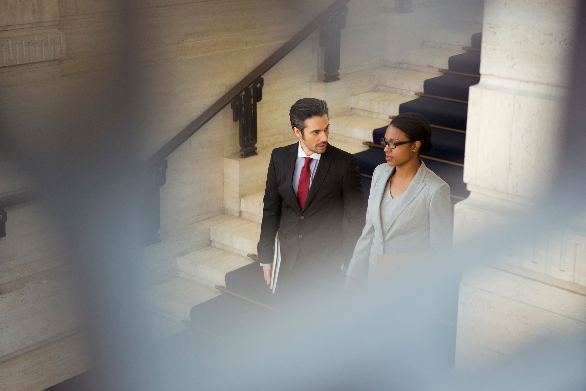 Two businesspeople walking up stone steps, one in a dark suit and red tie, the other in a light blazer and sunglasses