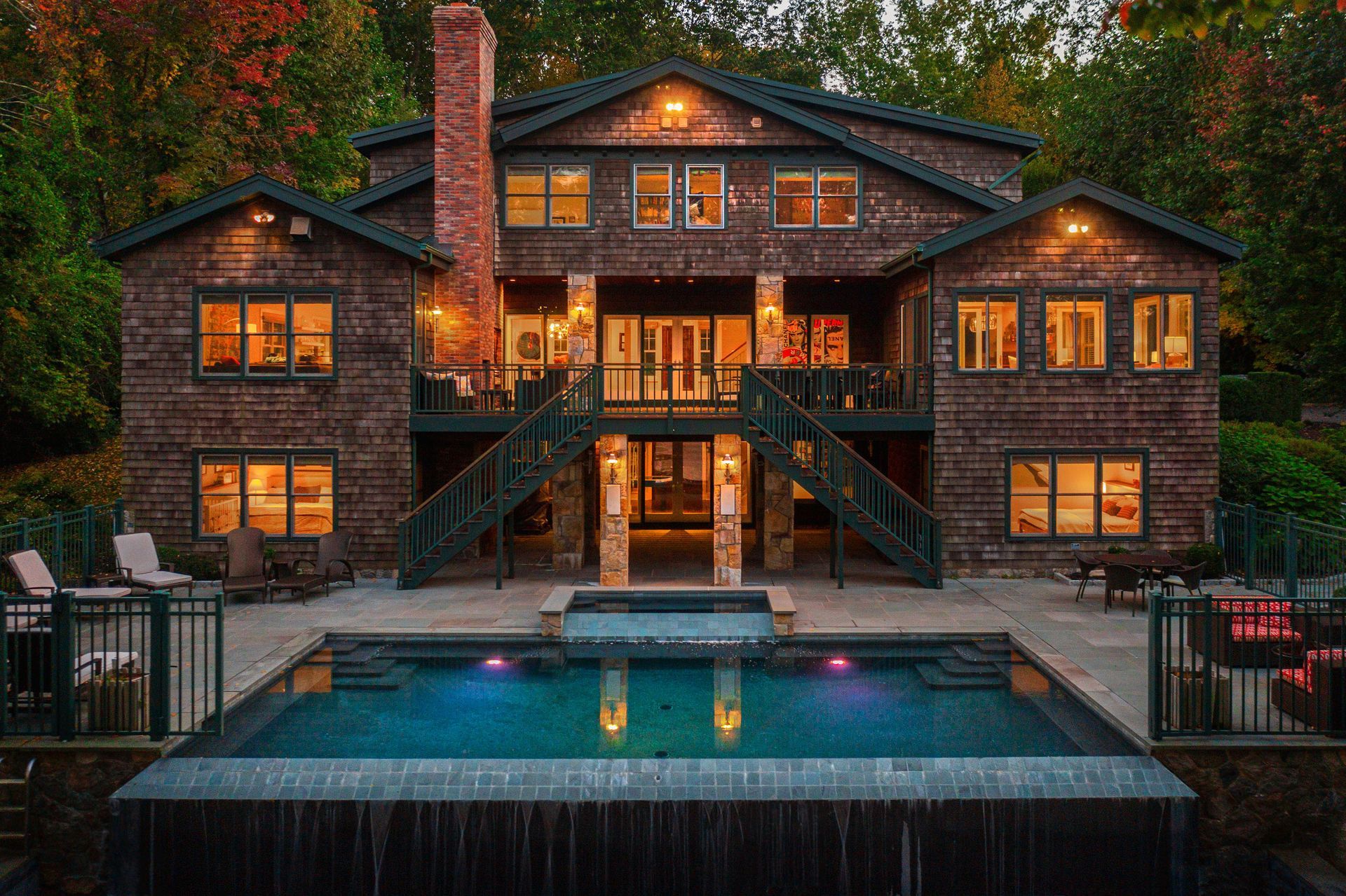 A multi-story stone home with an illuminated deck, staircase, and infinity pool at dusk, surrounded by trees.