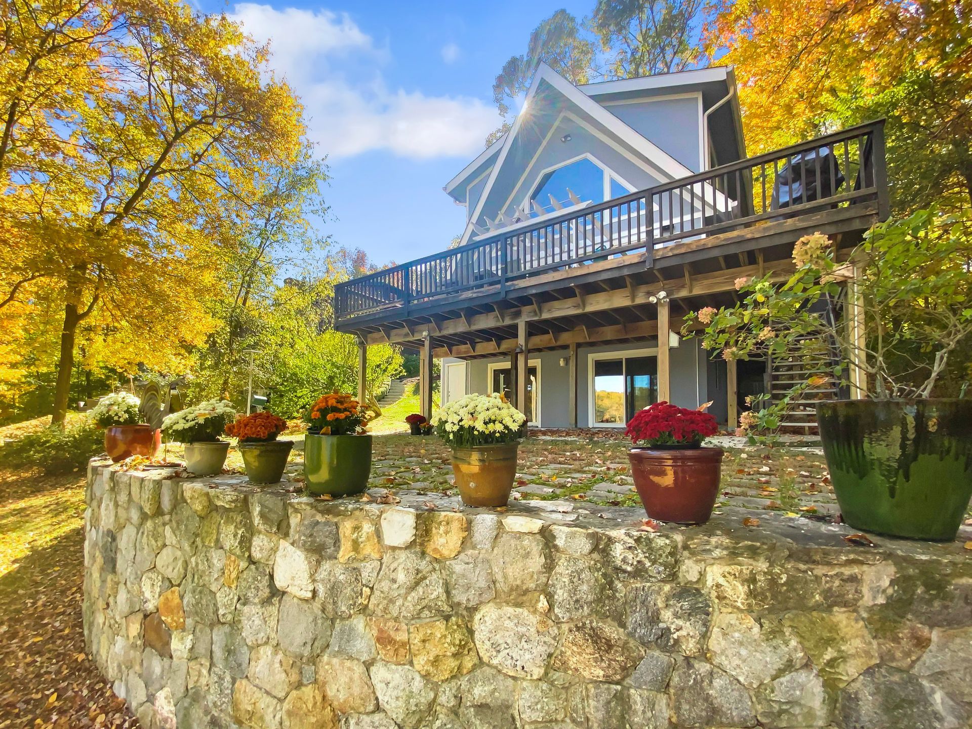 A light blue house with a wooden deck stands behind a stone retaining wall topped with several colorful potted flowers.