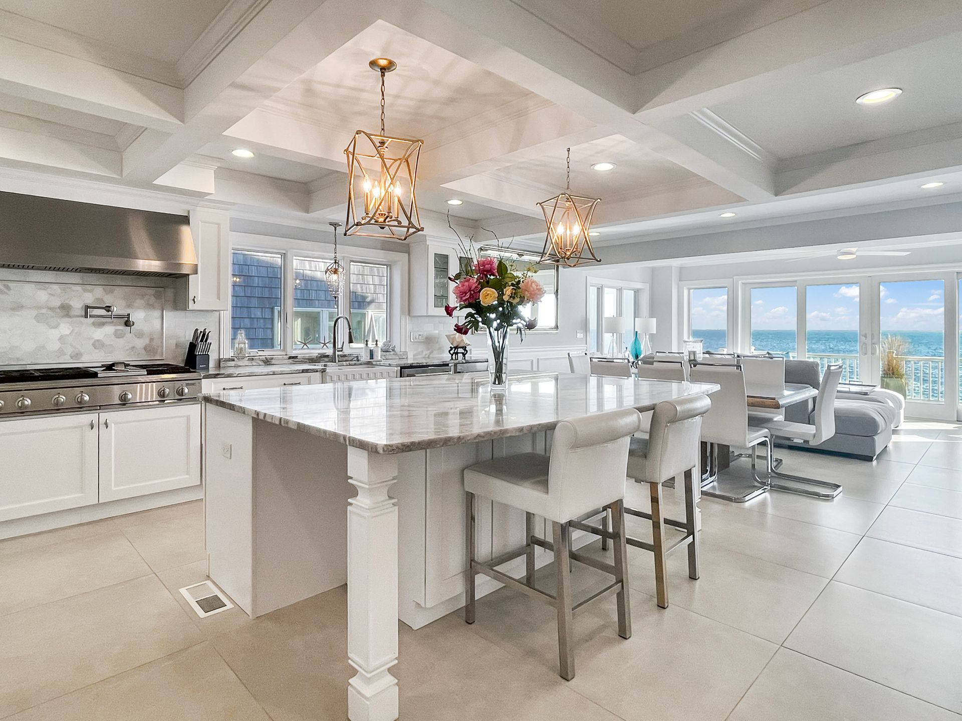 A bright, modern white kitchen with a large granite island, pendant lights, and a view of the ocean through large windows.