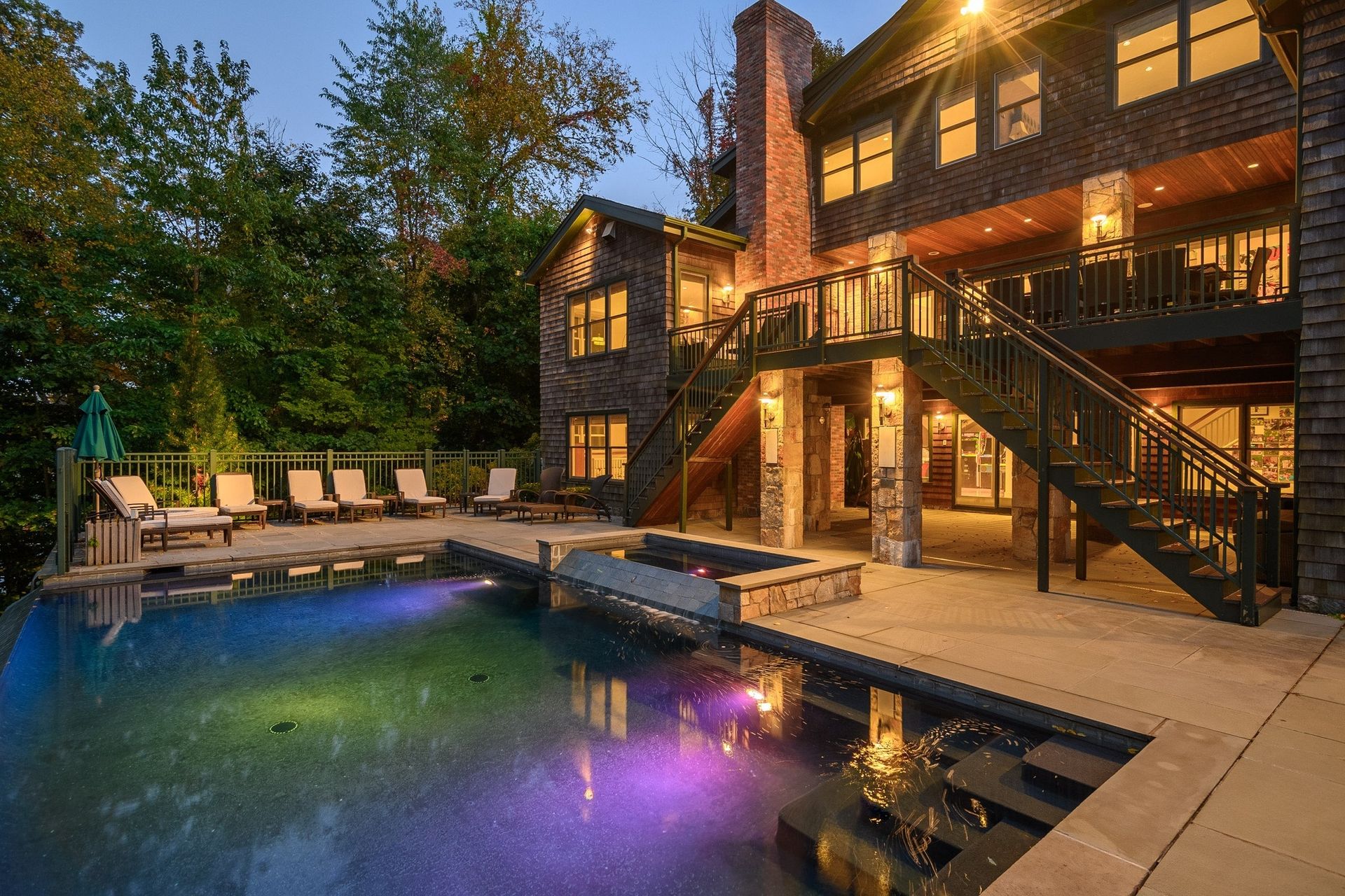 A multi-story house with a backyard pool, illuminated patio lights, and an outdoor staircase at twilight.
