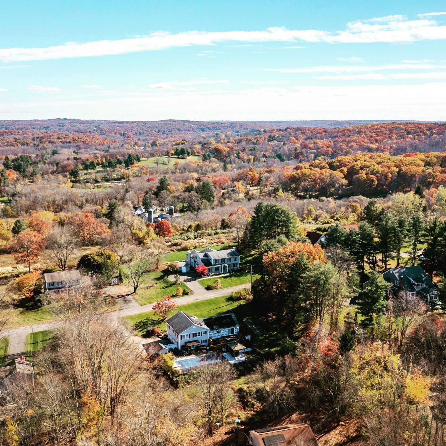 Aerial view of several houses nestled among colorful autumn trees on a rolling landscape under a bright, sunny sky.