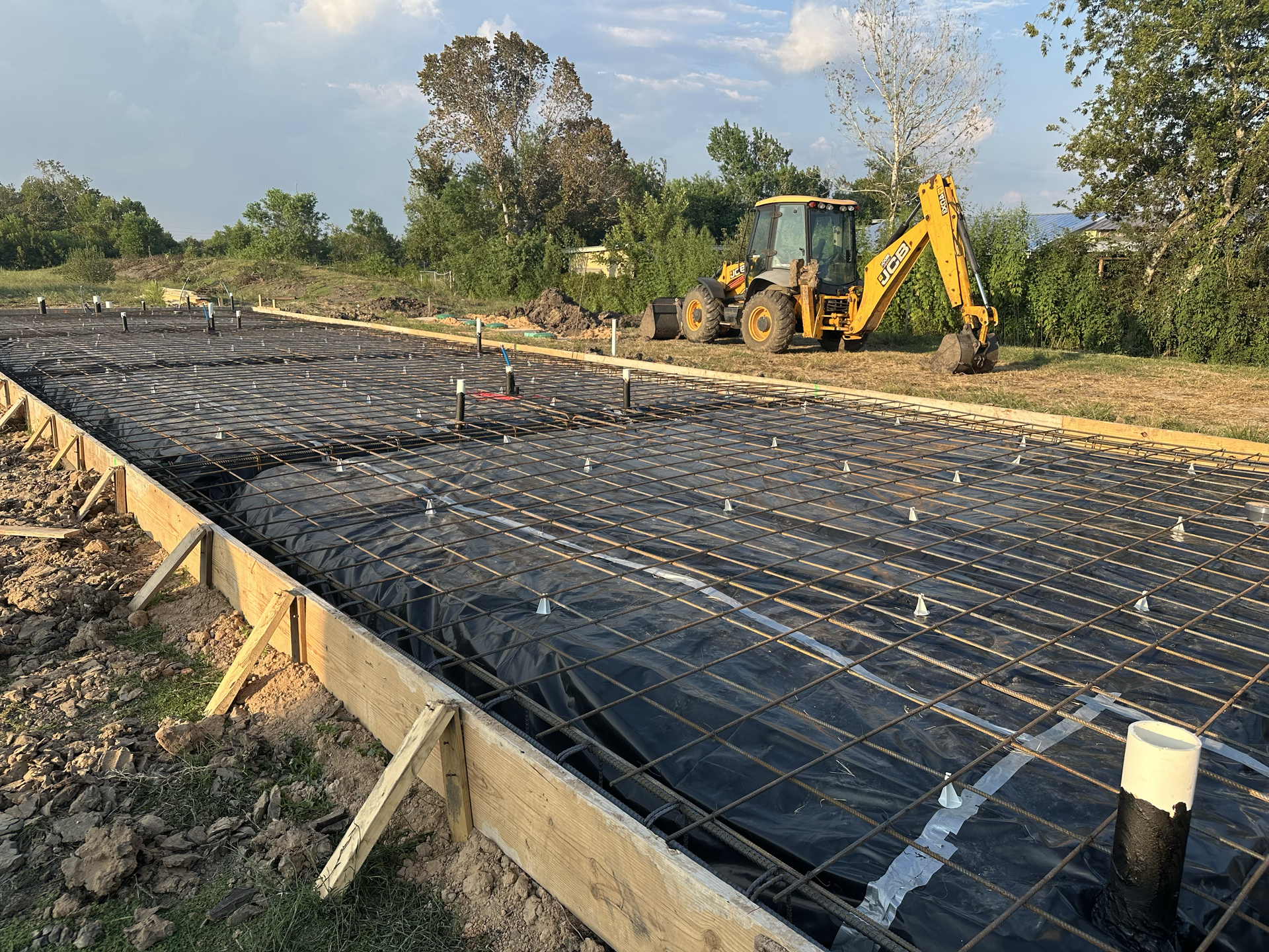 Construction site with wooden forms, rebar grid, black sheeting, and a yellow backhoe.
