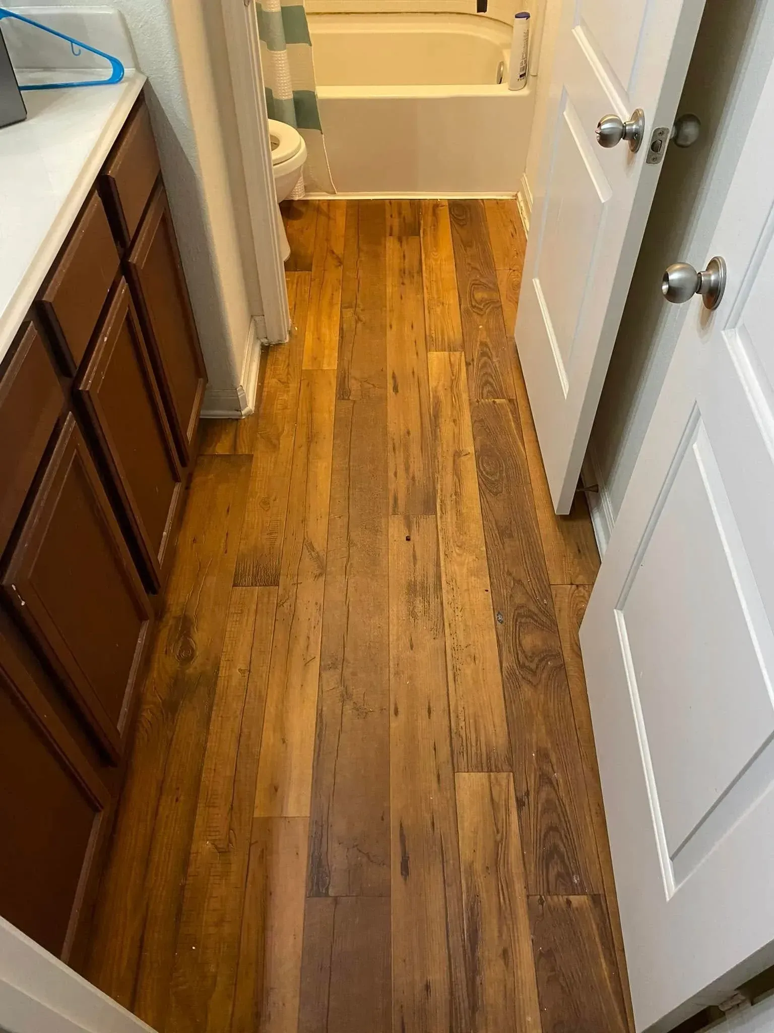 Bathroom with brown wood-look flooring, brown cabinets on left, white doors on right, and a bathtub in the back.