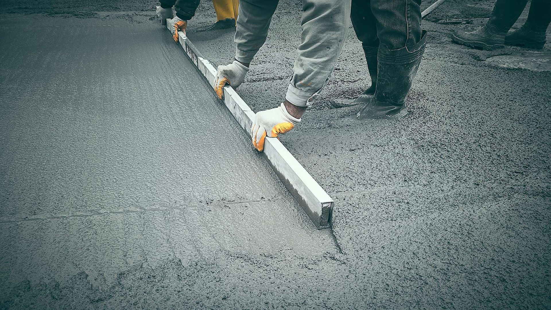 Workers leveling wet concrete with a long metal screed at a construction site.