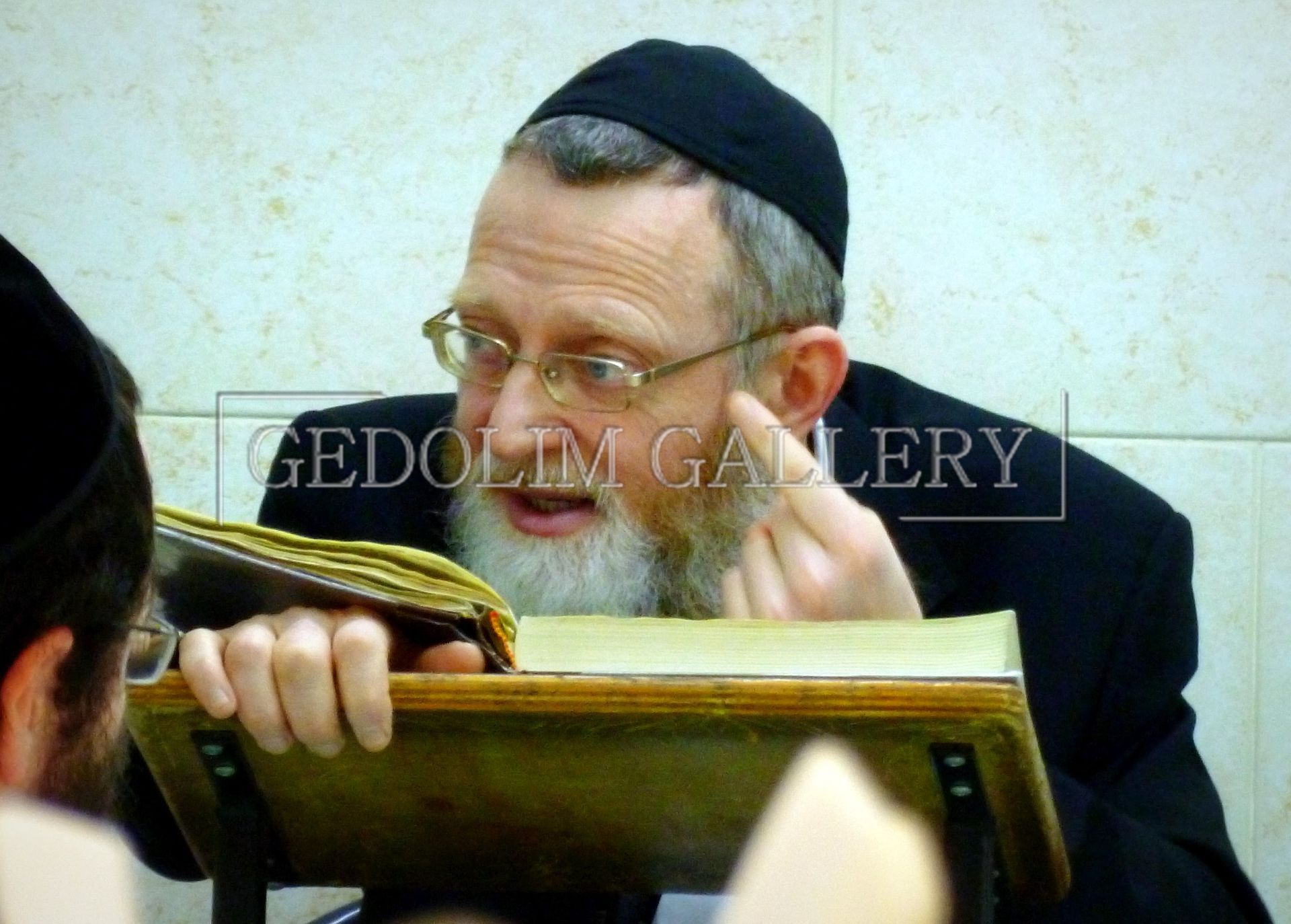 A man with glasses and a beard is reading a book in front of a wall that says gedolin gallery
