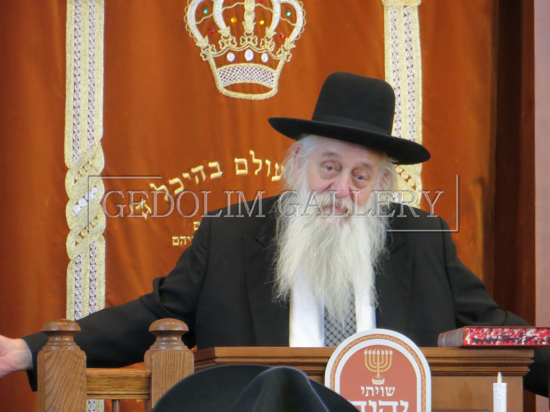 A man with a beard wearing a black hat stands in front of a sign that says gedolin gallery