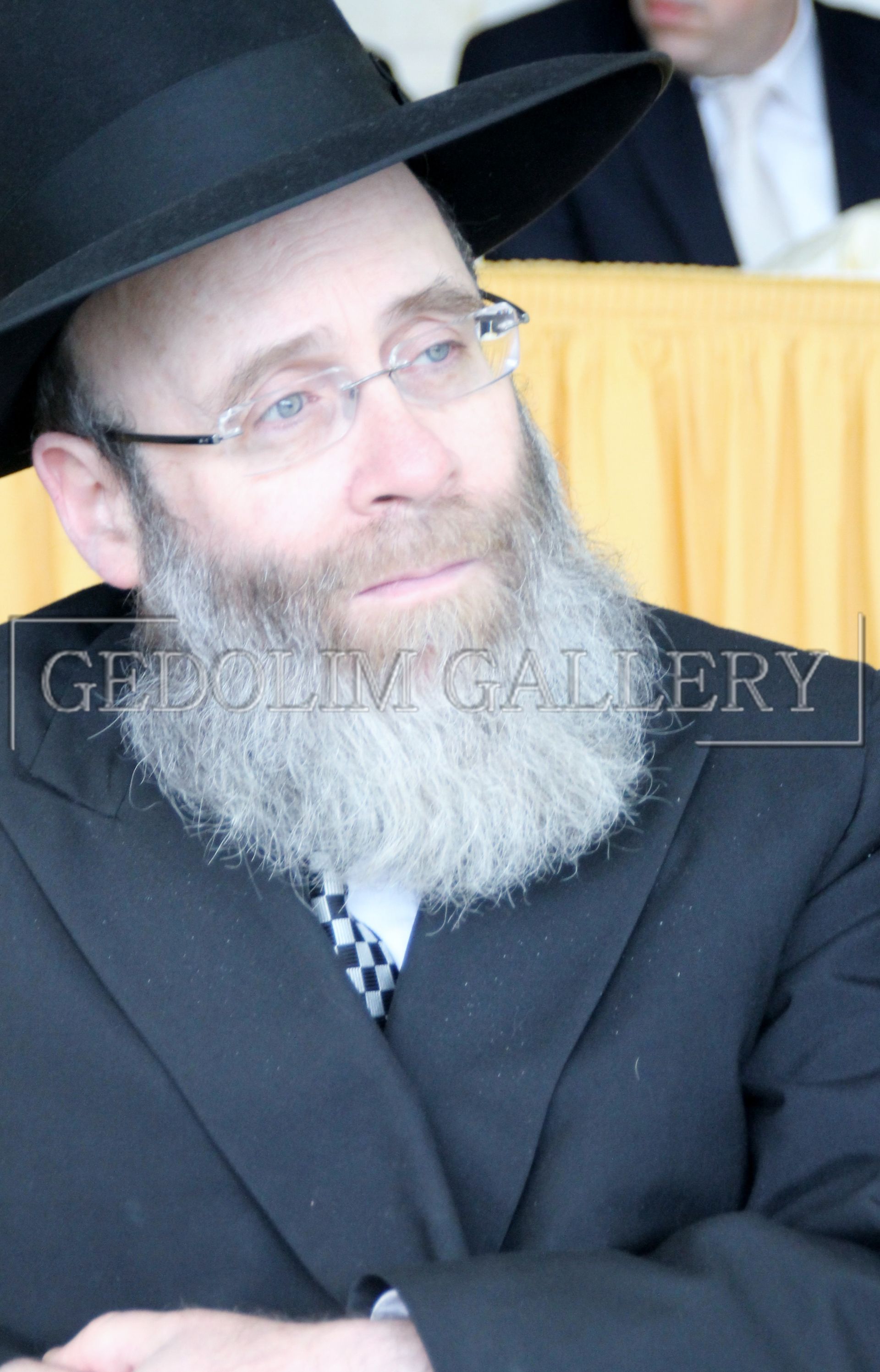 A man with a beard wearing glasses and a black hat is sitting in front of a yellow curtain with the word gallery on it