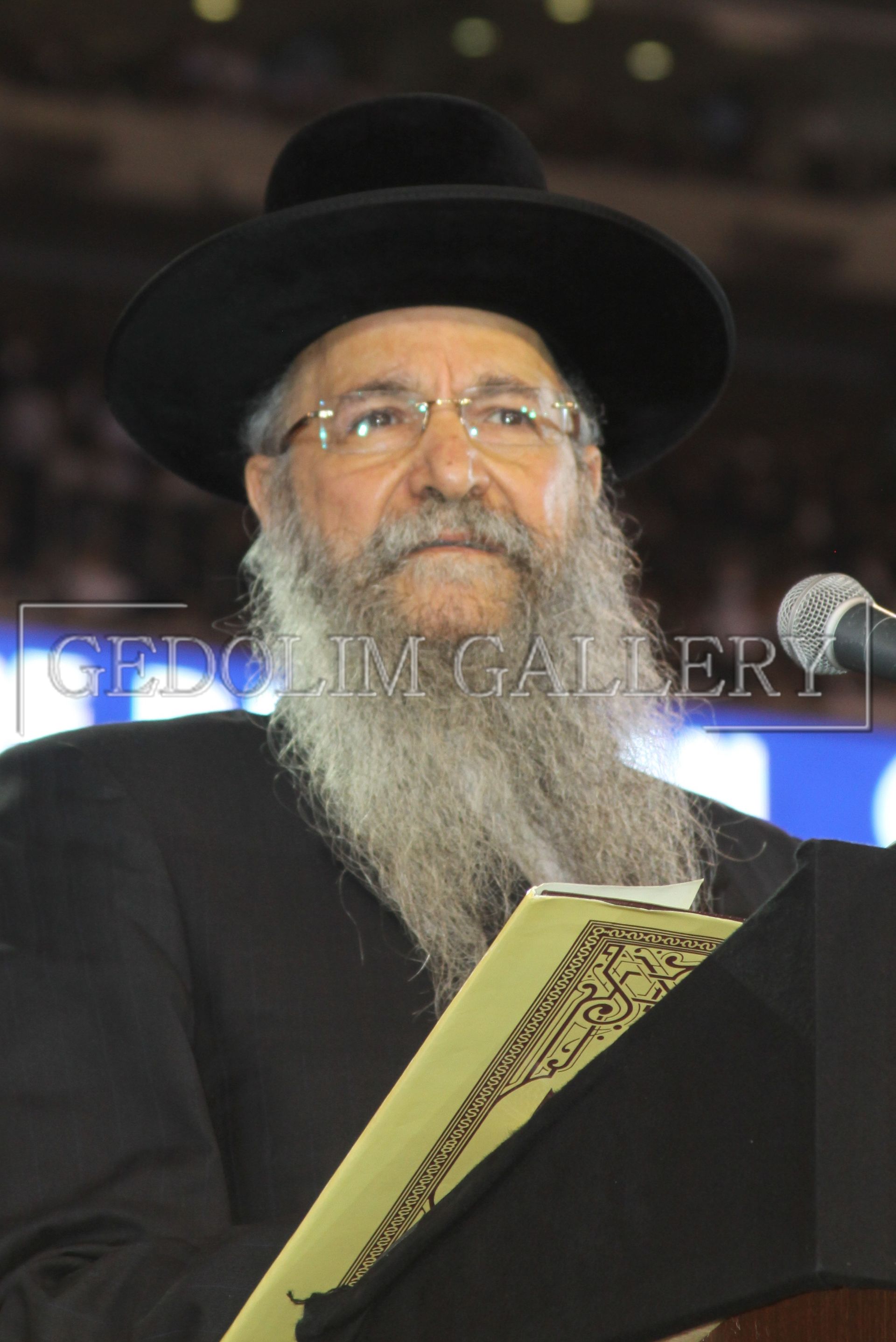 A man with a beard wearing a hat and glasses stands at a podium