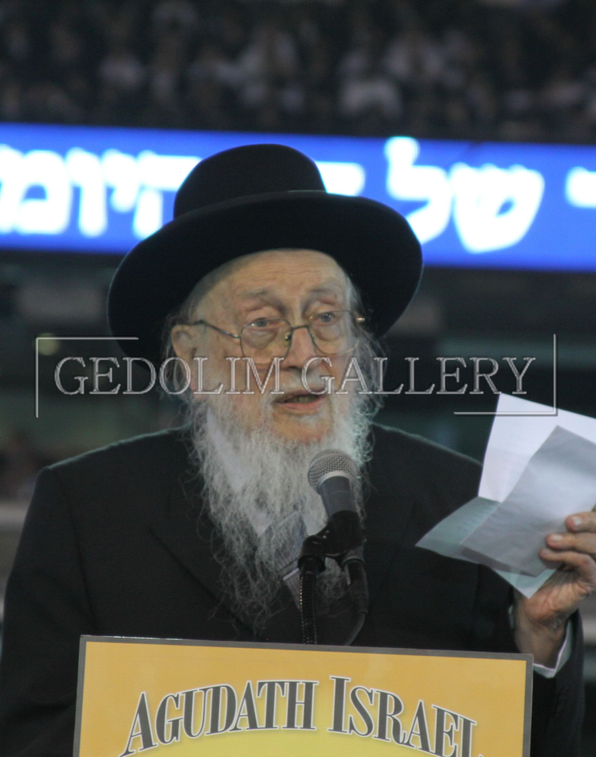 A man speaking into a microphone behind a sign that says agudath israel
