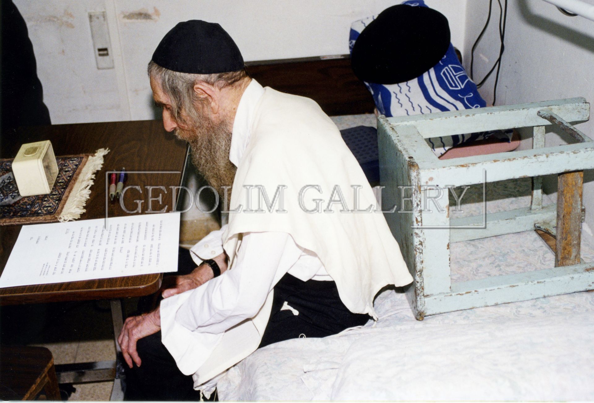 A man with a beard is sitting on a bed in front of a table with the word getolim gallery written on it