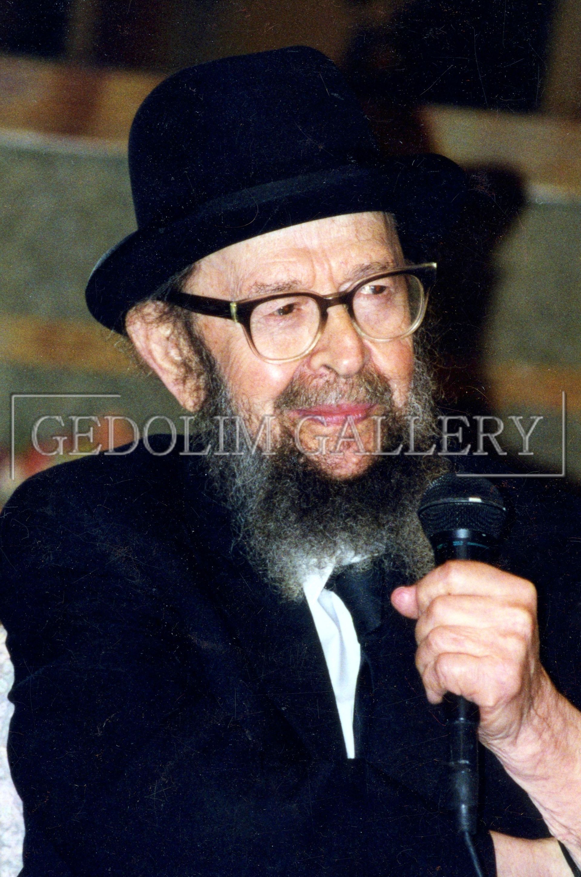 A man with a beard wearing a hat and tie is holding a microphone.