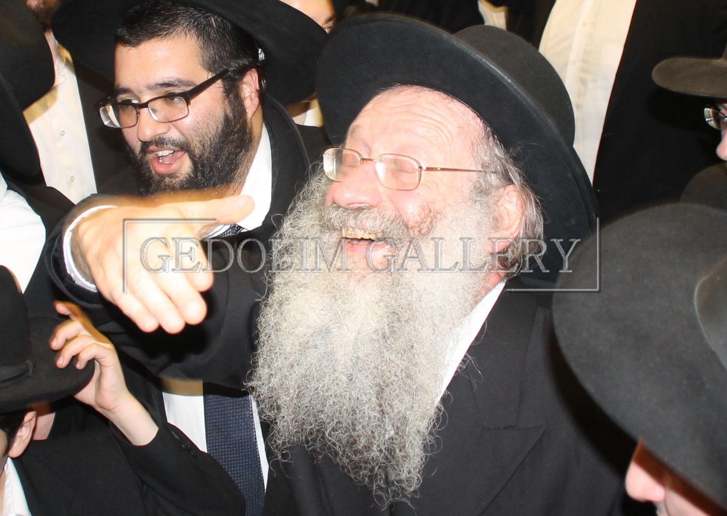 A man with a beard wearing glasses and a hat is surrounded by other men and the caption reads gedolin gallery
