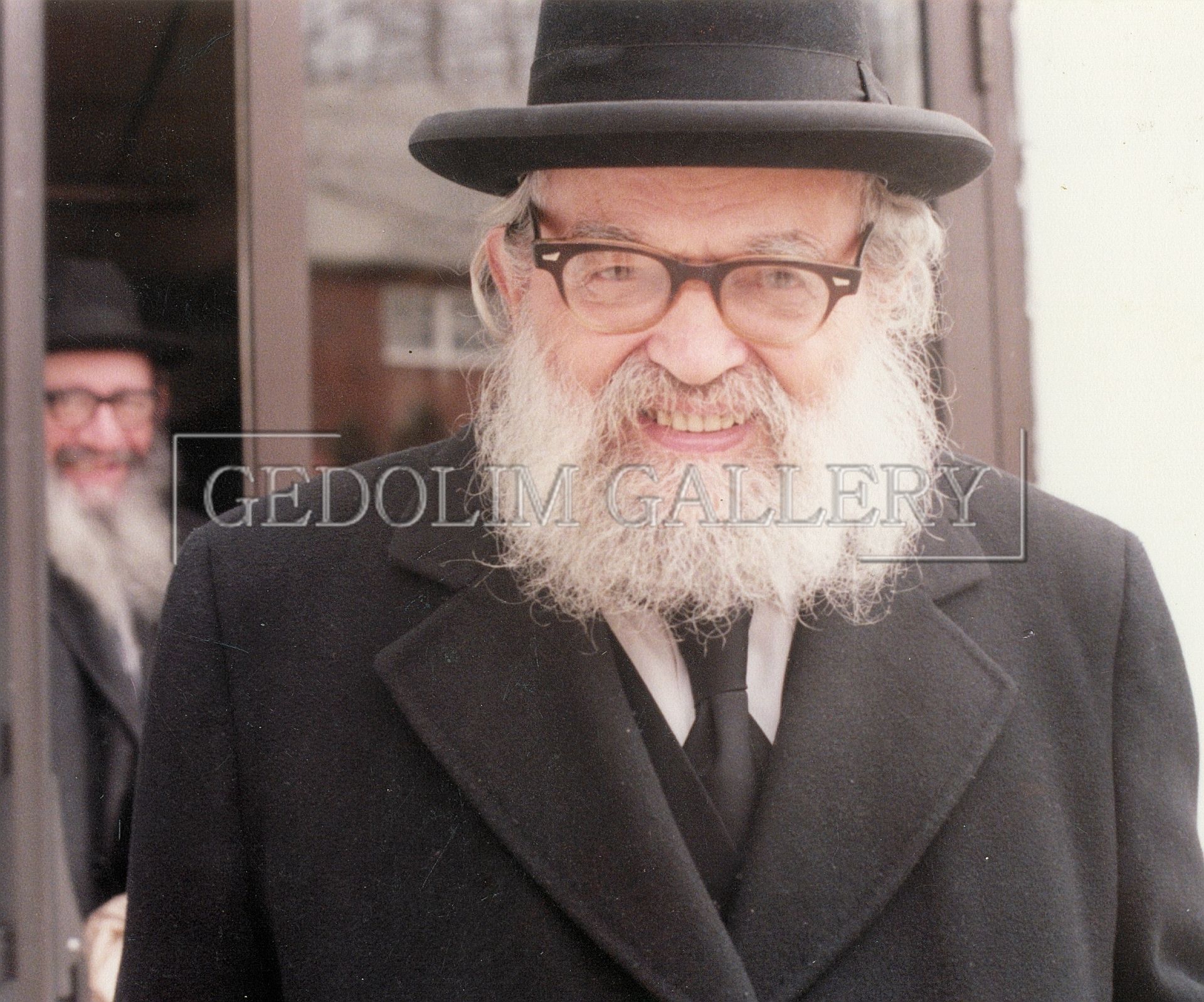 A man with a beard wearing glasses and a hat is standing in front of a gedolim gallery sign