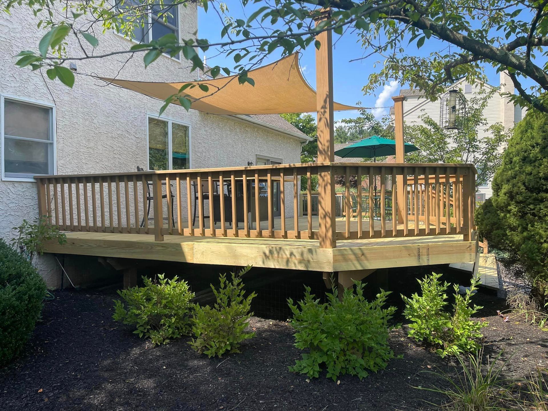 wood deck with railing, coming off the back of a stucco home. photo includes landscaping around bottom of deck and a sun awning above.