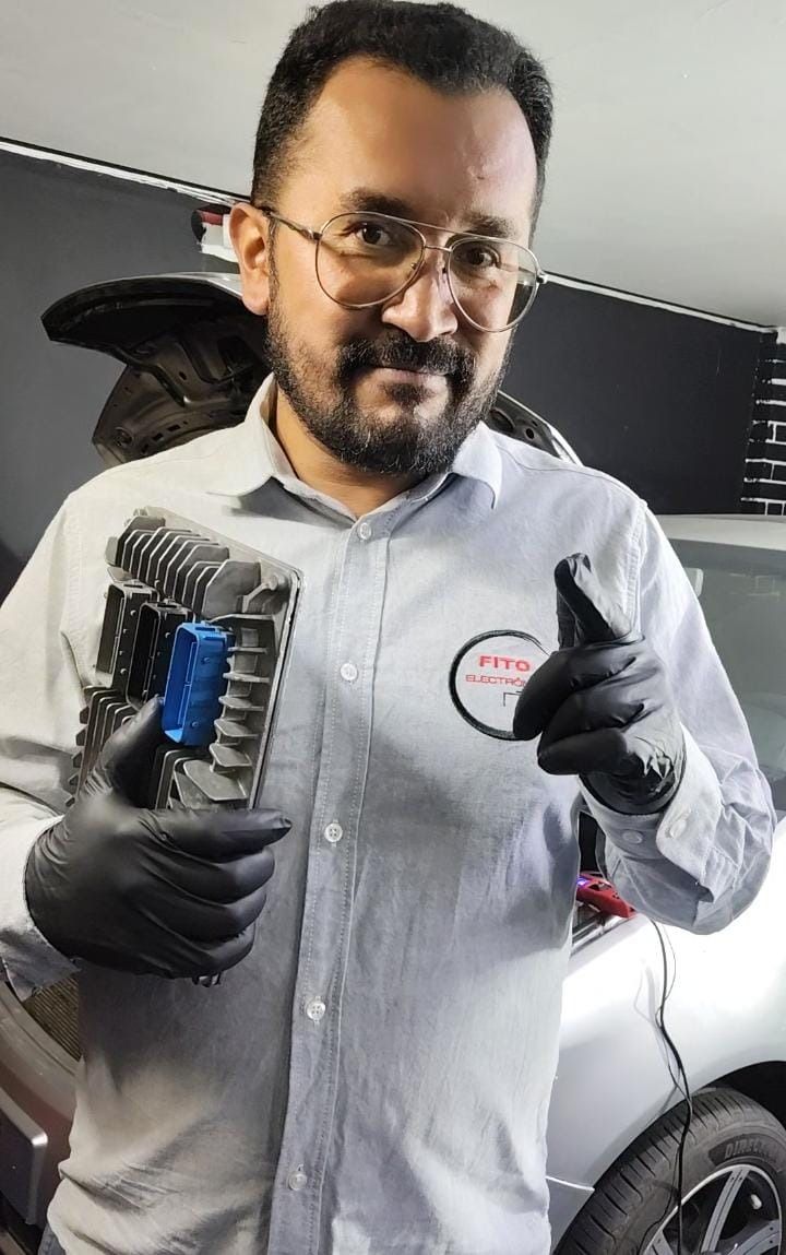 Técnico con camisa gris y guantes negros, sosteniendo una herramienta, de pie junto a un coche en un taller.